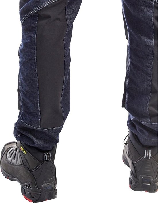 Detail van de zwarte versteviging op de onderpijp van een donkerblauwe werkspijkerbroek met schoenen.