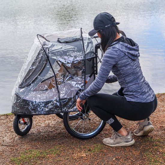 Vrouw met petje met regenhoes over een grijze hondenbuggy. Een hond kijkt uit het gaas.
