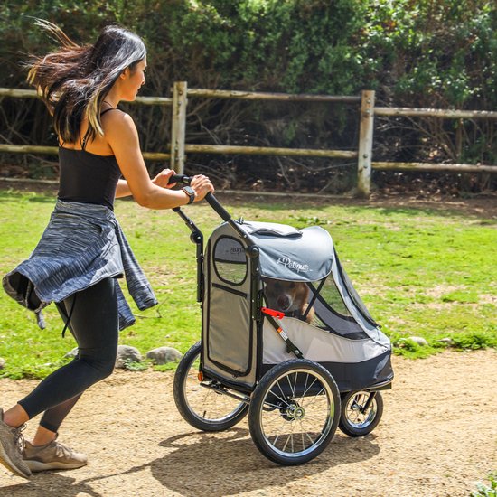 Vrouw met donker haar in sportkleding duwt een grijze hondenbuggy met grote wielen door een bosrijk gebied.
