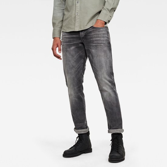 Man in grijze G-Star 3301 regular tapered jeans met opgerolde zoom, grijs overhemd en zwarte laarzen.