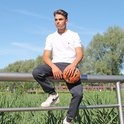 Man zit op een hek met een bal, draagt een donkere joggingbroek en witte t-shirt.