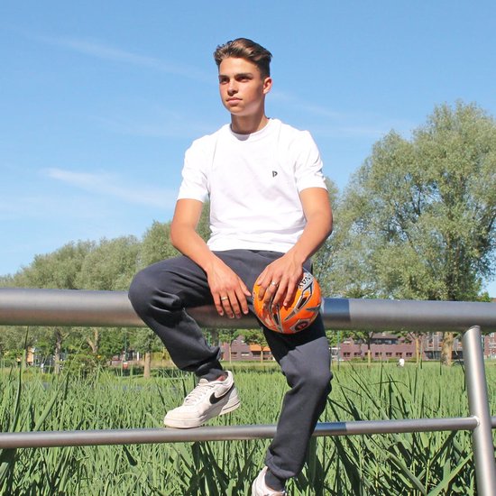 Man zit op een hek met een bal, draagt een donkere joggingbroek en witte t-shirt.