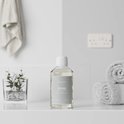 Fles Steamstory wasparfum 'Pearl' op een plank met handdoeken en een plant.