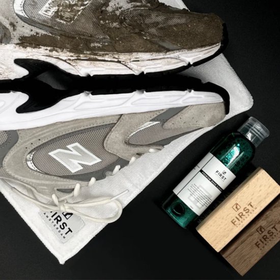 Fles met groene schoenreiniger, naast een schone en een vuile sneaker op een witte handdoek. Twee houten borstels met logo.