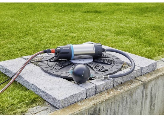 Zilverkleurige dompelpomp met zwarte details, aangesloten op een grof filter met een zwarte bal, geplaatst op een stenen tegel in gras.