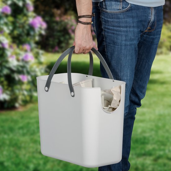 Man draagt een witte afvalbak met grijze handvatten, gevuld met papier, in een tuin.