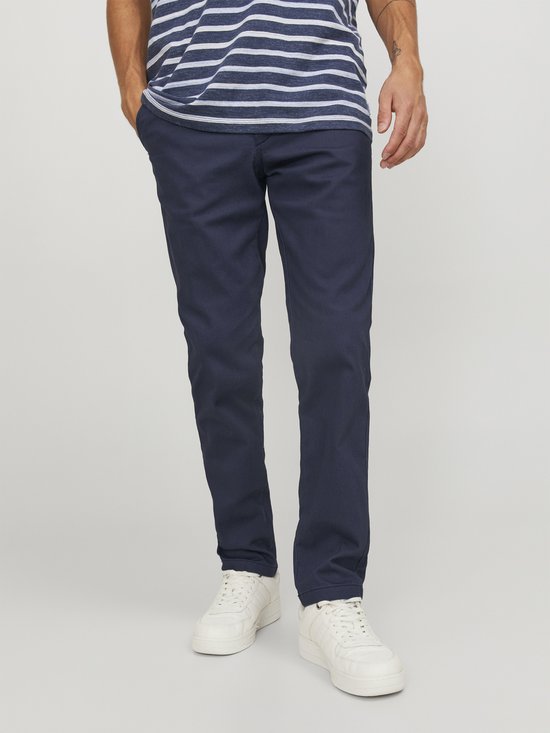 Man in gestreept t-shirt en marineblauwe chino broek, staand met handen in zakken.