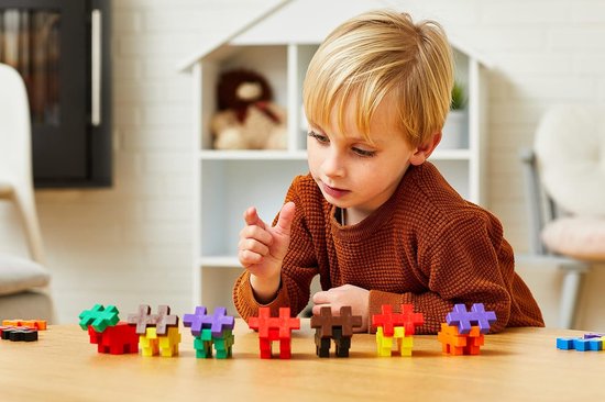 Een jongen met blond haar bekijkt Plus-Plus constructies van groene, rode, bruine en gele blokjes op een houten tafel.