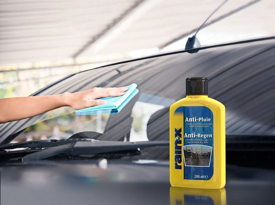 Gele fles Rain-X Anti-Pluie/Anti-Regen naast de voorruit van een auto, hand maakt schoon.