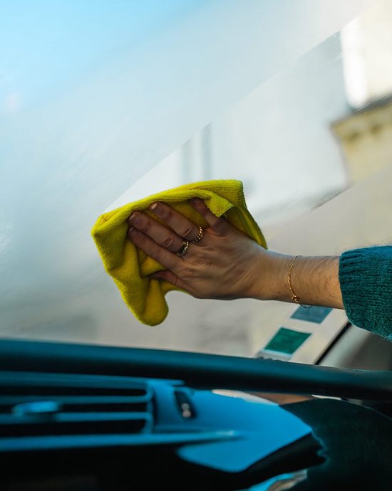 Een hand met ringen gebruikt een gele doek om een beslagen autoruit schoon te maken.