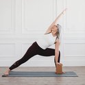 Femme aux longs cheveux gris en legging violet et haut blanc, en posture de yoga avec un bloc de yoga en liège marron.