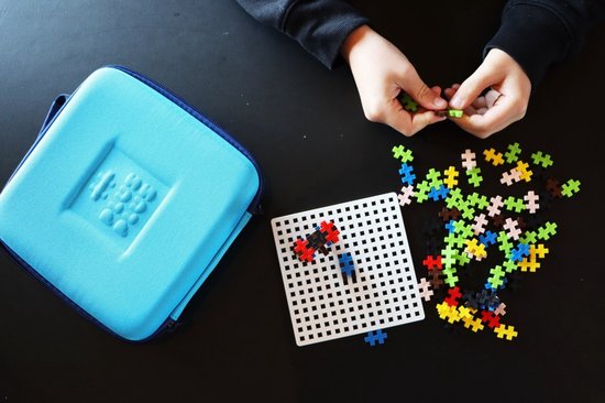Een kinderhand bouwt aan een auto met Plus-Plus speelgoedblokjes op een wit pegboard, naast een turquoise koffer en verspreide blokjes.