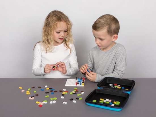 Twee kinderen, een meisje met blonde krullen en een jongen, spelen met Plus-Plus bouwstenen aan tafel. Een open koffer met stenen staat ernaast.