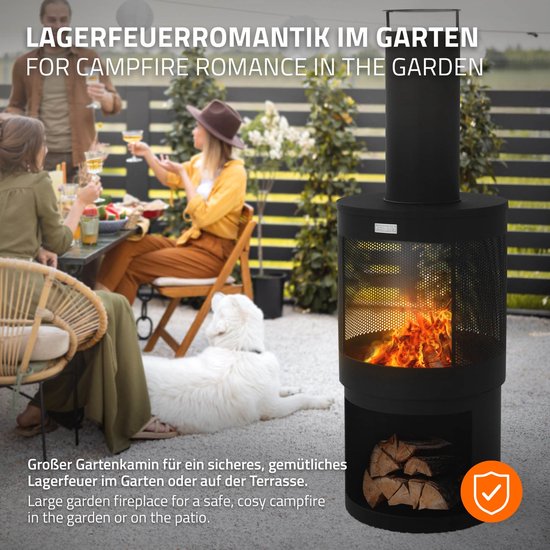 Zwarte tuinhaard met vlammen en hout, in een tuin met mensen, hond en verlichting.