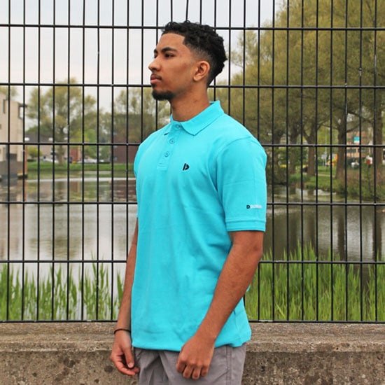 Man staat buiten in turquoise polo met korte mouwen, grijze broek en Donnay logo.