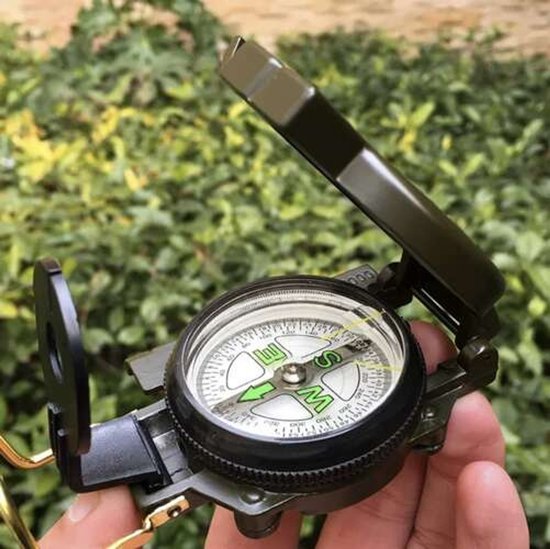 Een hand houdt een groen, inklapbaar militair kompas vast met de wijzerplaat en kompasnaald zichtbaar.