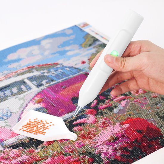Main tenant un stylo blanc électrique pour peinture diamant au-dessus d'une œuvre d'art colorée avec un plateau plein de diamants orange.