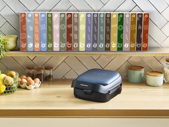 Een gesloten, donkergrijs Tefal wafelijzer staat op een houten aanrecht met diverse Tefal Snack Collection dozen op de achtergrond.