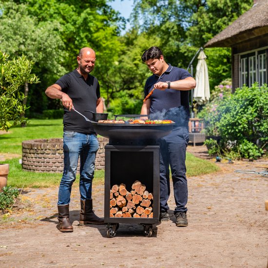 Twee mannen barbecueën bij een zwarte, ronde grill met houtopslag. De een draait vlees, de ander prikt een spies.