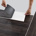 Handen die de beschermlaag van een donkerbruine PVC vloerplank met houtnerf aftrekken.