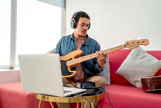 Zwarte RØDE AI-1 audio interface op een salontafel, aangesloten op een laptop, terwijl een man basgitaar speelt met koptelefoon.