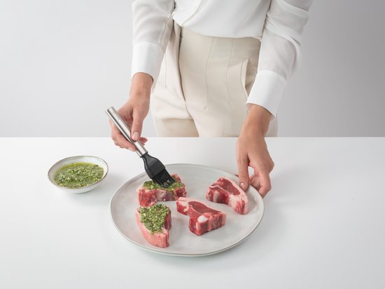 Mains badigeonnant des côtelettes d'agneau avec un mélange vert à l'aide d'un petit pinceau de pâtisserie argenté à poils noirs.