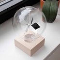 Glazen bol met houten voet, bevat een staaf met aan weerszijden schijven, deels zilverkleurig en deels zwart, bij een raam met planten.