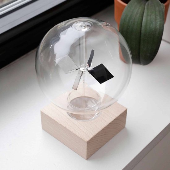 Glazen bol met houten voet, bevat een staaf met aan weerszijden schijven, deels zilverkleurig en deels zwart, bij een raam met planten.