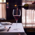 Iemand schenkt rode wijn in een glas op een gedekte tafel met witte tafelkleden en servetten.