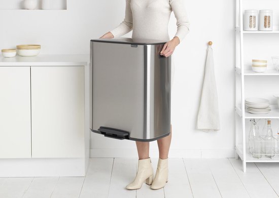 Vrouw met beige laarsjes tilt een mat stalen Brabantia prullenbak op.
