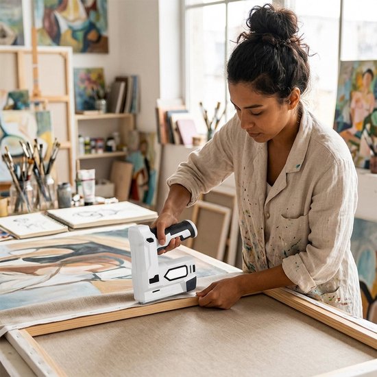 Vrouw in een lichtgekleurd hemd gebruikt een wit-zwart nietpistool op een canvasdoek in een atelier.