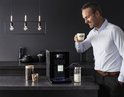 Man met lichtblauwe blouse geniet van een latte macchiato naast de zwarte Krups Intuition Preference machine.