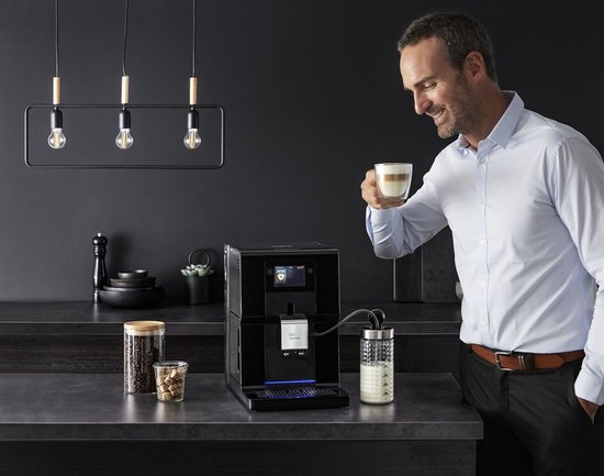 Man met lichtblauwe blouse geniet van een latte macchiato naast de zwarte Krups Intuition Preference machine.