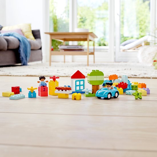 LEGO Duplo speelgoed op een houten vloer. Een blauwe auto, een huisje, een boom, twee poppetjes, blokken en een podium met de cijfers 1, 2, 3.