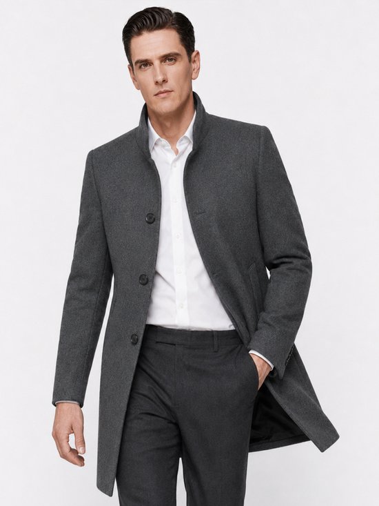 Man in grijze wollen jas met staande kraag, drie knopen, witte blouse, hand in de zak.