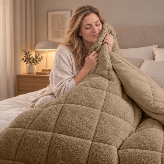 Een vrouw met gesloten ogen omhelst een dik, taupe teddy dekbed dat op een bed ligt.