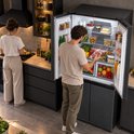 Man pakt een stuk vlees uit een open, donkergrijze Heinner Amerikaanse koelkast in een keuken, terwijl een vrouw kookt.