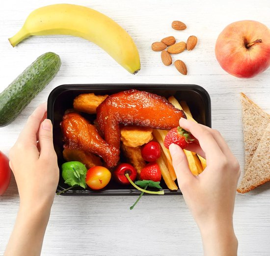Handen vullen een zwarte voedselcontainer met kip, frietjes, fruit en groenten, naast een banaan, appel, komkommer en amandelen.
