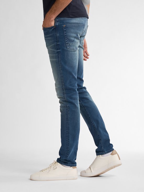Model draagt een Petrol Industries slim fit jeans in blauwe vintage wassing met 6 zakken, gecombineerd met witte sneakers.