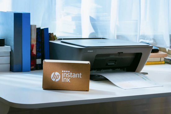 Zwarte HP DeskJet printer op bureau naast een doos met HP Instant Ink-logo.