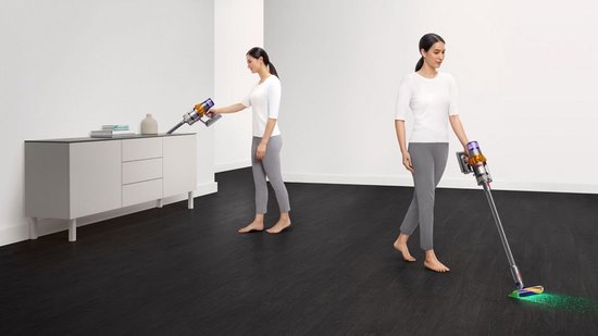 Een vrouw met een witte top en grijze broek gebruikt een Dyson V15s steelstofzuiger met dweilfunctie op een donkere vloer.