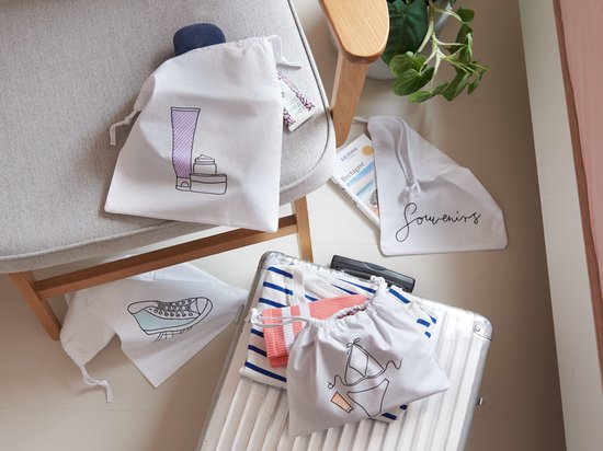 Witte stoffen zakjes met tekeningen van cosmetica, een sneaker en 'Souvenirs'. Een deel van de zakjes ligt op een koffer.