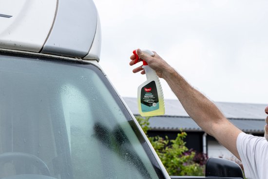 Een hand spuit de gele vloeistof uit een fles met rode trigger op een natte voorruit. Het etiket 'INSECTFREE' is zichtbaar.
