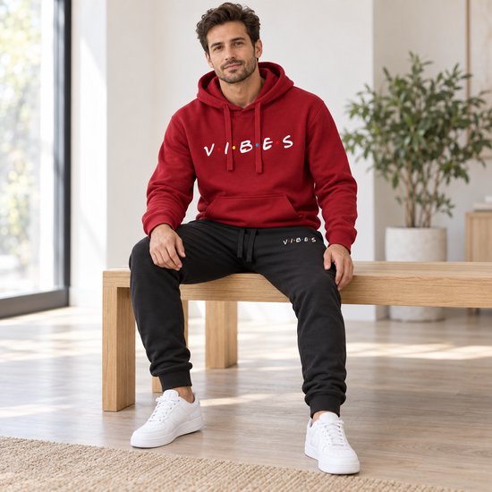 Man met bordeauxrode hoodie met 'VIBES'-opdruk en zwarte joggingbroek met 'VIBES'-opdruk, zittend op een houten bank.