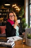 Vrouw met bril en een roze sjaal om haar nek drinkt koffie bij een laptop.