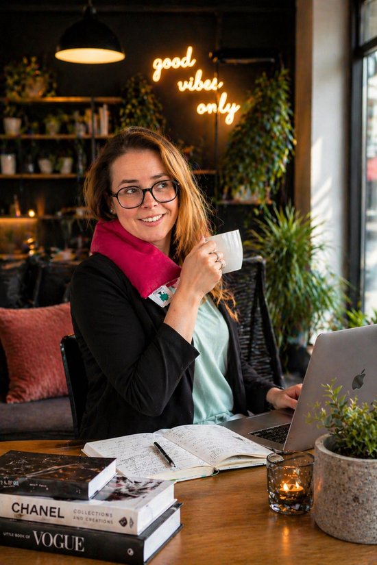 Vrouw met bril en een roze sjaal om haar nek drinkt koffie bij een laptop.