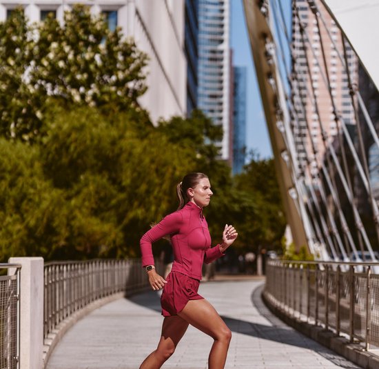 Vrouw in bordeauxrode sportkleding jogt op een stadsbrug met Fitbit Edge om de pols.