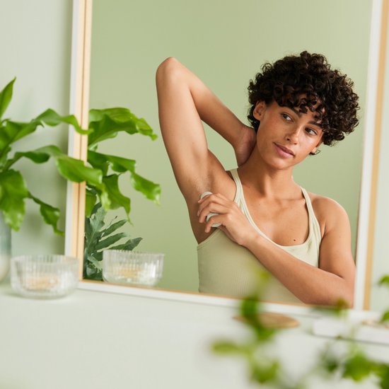 Vrouw met krullend haar in een lichte tanktop brengt WELEDA deodorant aan onder haar arm in een badkamer met planten.