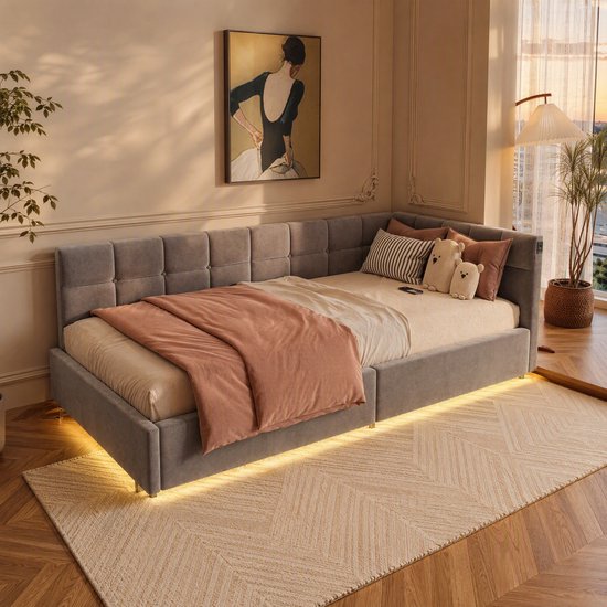 Grijs fluwelen hoekbed met openklapbare matras, roze plaid en decoratieve kussens, met gele LED-verlichting.