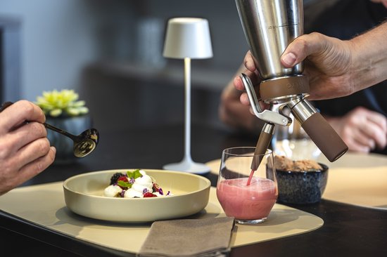 Roze drankje wordt uit een roestvrijstalen nitro-sifon in een glas geschonken naast een dessertbord.